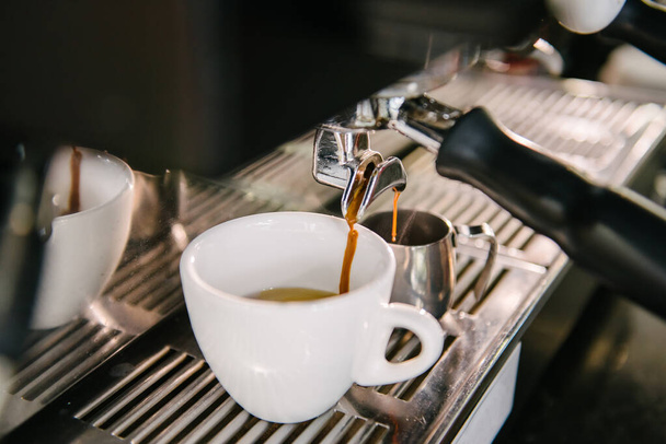 En la taza blanca que está en la máquina de café, el café fresco con espuma fluye fuera del soporte - Foto, Imagen