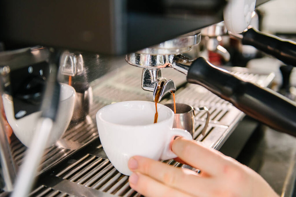 The barista is holding a white cup in the coffee machine. Fresh coffee with foam flows out of the holder - 写真・画像