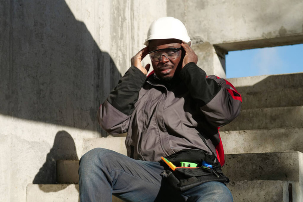 Afrikai amerikai munkás ül a lépcsőn az építkezésen. Nem érzi jól magát és fáj a feje, megérinti a fejét. - Fotó, kép