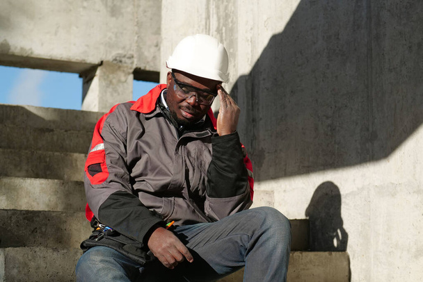 African american workman sits on the stairs on construction site. He is not feeling well and has headache, he touches his head - Foto, Imagen