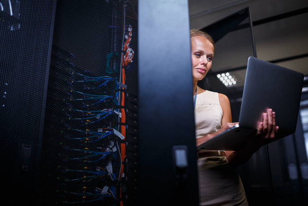 IT engineer standing amid working server racks doing routine maintenance check and diagnostics using laptop computer (color toned image) - Fotó, kép
