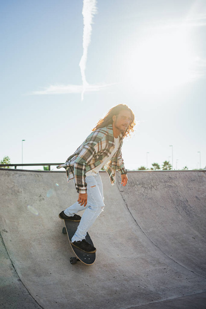 Foto retroilluminata di un uomo dai capelli lunghi che scende da una rampa con un surfskate in uno skate park - Foto, immagini