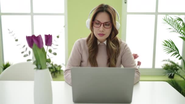 Young beautiful hispanic woman using laptop and headphones sitting on table at home - Filmmaterial, Video