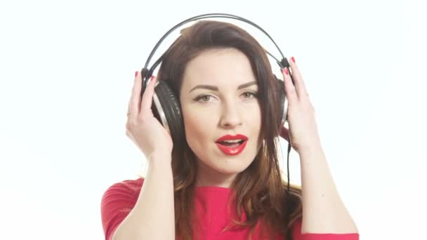 wunderschönes Mädchen in Rot, das mit der Musik in großen Kopfhörern schunkelt, die Kamera bemerkt und isoliert auf weißem Hintergrund Nahaufnahme scheut - Filmmaterial, Video