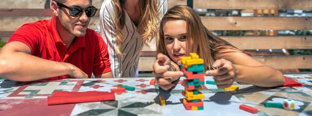 Mujer joven concentrada empujando pieza de juego de jenga junto a sus amigos sonrientes en la azotea en una fiesta de verano - Foto, Imagen