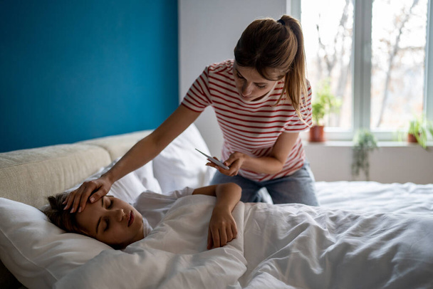 Worried caucasian mother measuring temperature of sick teen daughter lying in bed at home, caring mom parent holding thermometer touching head of unhealthy preteen child. Children and colds - Foto, Imagen