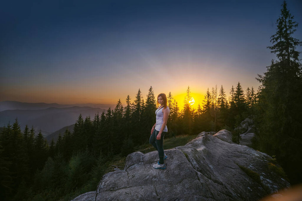 A lány a sziklán, a szikla tetején. Naplemente egy nyári napon. A fenyőfák csúcsa. Az aktív pihenés, túrázás és utazás fogalma. Gyönyörű kilátás a Kárpátokra, Ukrajna. - Fotó, kép