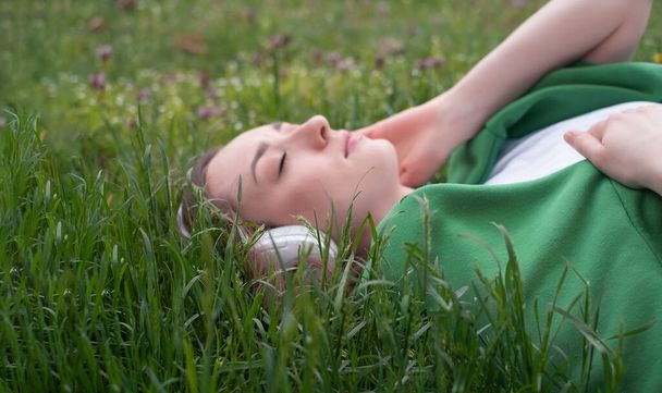 Genç bir kız kulaklıkla müzik dinliyor, parktaki yeşil çimenlerde yatıyor, gülümsüyor ve dilini beyaz tişört ve yeşil kazakla gösteriyor, yaz havası yaşam tarzı olarak. - Fotoğraf, Görsel