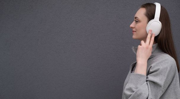 Carefree atraktivní brunetka dívka, oblečená v šedém svetru, poslech hudby na sluchátkách, stojící na šedém pozadí - Fotografie, Obrázek