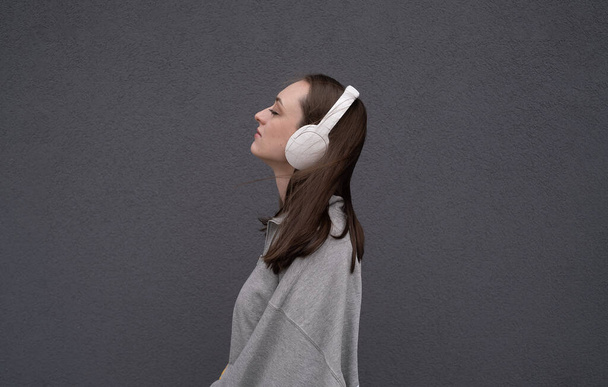 Chica morena atractiva despreocupada, vestida con un suéter gris, escuchando música en los auriculares, de pie sobre un fondo gris - Foto, imagen