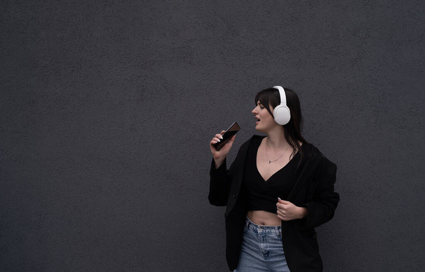 Una joven adolescente escucha música con auriculares, vestida con una chaqueta negra y jeans, baila con la música y canta canciones en voz alta - Foto, Imagen