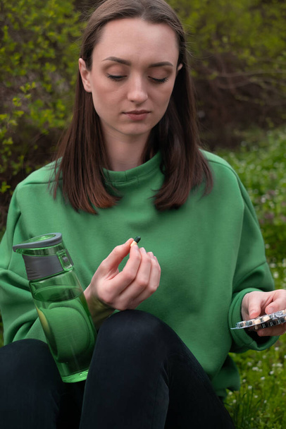 het meisje houdt vitaminen in haar hand, en een fles in het voorjaar in het bos, gekleed in een groene jas - Foto, afbeelding