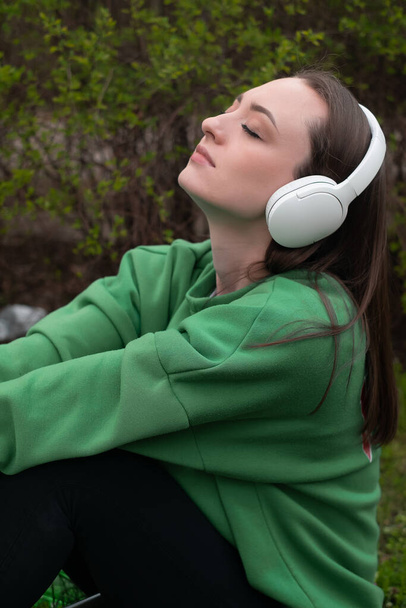Mladá žena sedí v zelené jarní trávě a baví se hudbou v parku, poslouchá ve sluchátkách se zavřenýma očima a blaženým úsměvem, oblečená do zelené trička - Fotografie, Obrázek