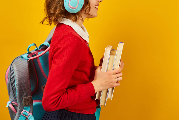 Estudante irreconhecível com mochila, fone de ouvido e livros na mão de volta à escola no fundo do estúdio amarelo, menina adolescente em uniforme escolar. - Foto, Imagem