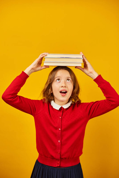 Retrato de adolescente feliz colegiala en uniforme sosteniendo libros en la cabeza en el fondo del estudio amarillo, divertirse vacaciones, concepto de regreso a la escuela. - Foto, imagen