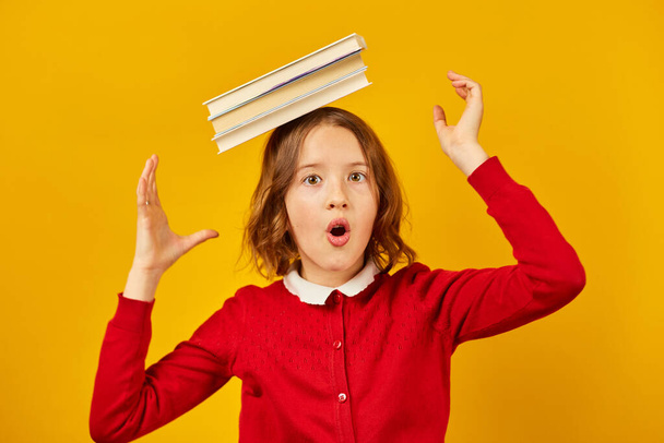 Портрет щасливої школярки-підлітка в уніформі, що тримає книги на голові на жовтому фоні студії, веселий відпочинок, концепція "Назад до школи"
. - Фото, зображення