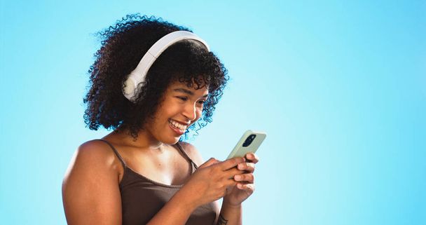 Black woman, phone and headphones isolated on blue background listening to music, social media video or funny meme. Happy gen z person laughing for audio tech, internet post and cellphone in studio. - 写真・画像