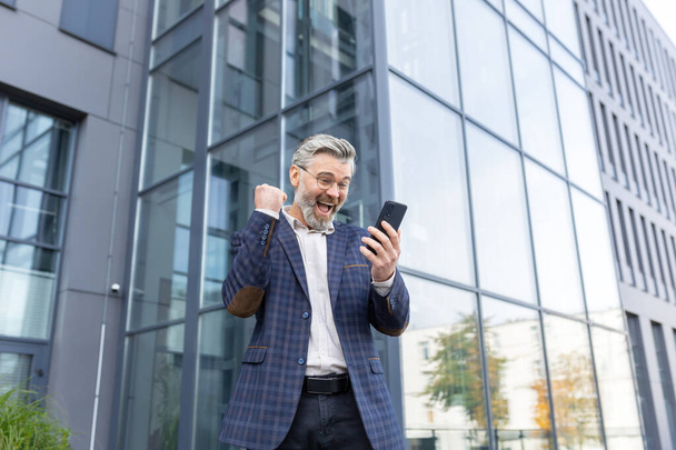 A senior man in a suit stands near an office center and is happy. Shows a victory gesture with his hand and shouts. He looks at his phone, reads a message, a letter, got a job, position, promotion. - 写真・画像