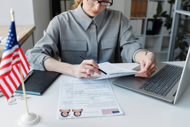 Hoge hoek schot van volwassen blanke vrouw dragen van grau shirt zitten aan het bureau in het werken met visa aanvraagformulieren - Foto, afbeelding