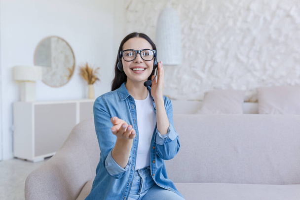 Beautiful young woman in headset sitting at home on sofa in front of camera and vlogging, recording blog, conducting webinar, growth and development trainings, pointing at camera, smiling. - Fotó, kép