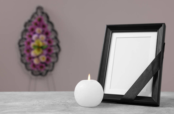 Photo frame with black ribbon, burning candle on table and wreath of plastic flowers near grey wall, space for text. Funeral attributes - 写真・画像