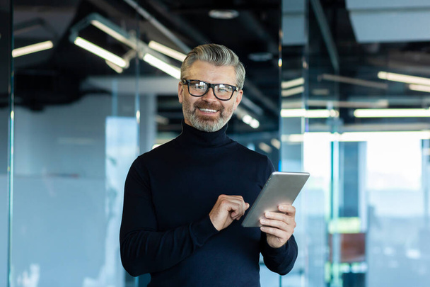 Portrait d'un sénior beau homme d'affaires aux cheveux gris dans des lunettes debout dans le bureau, en utilisant une tablette et en regardant l'appareil photo avec un sourire. - Photo, image
