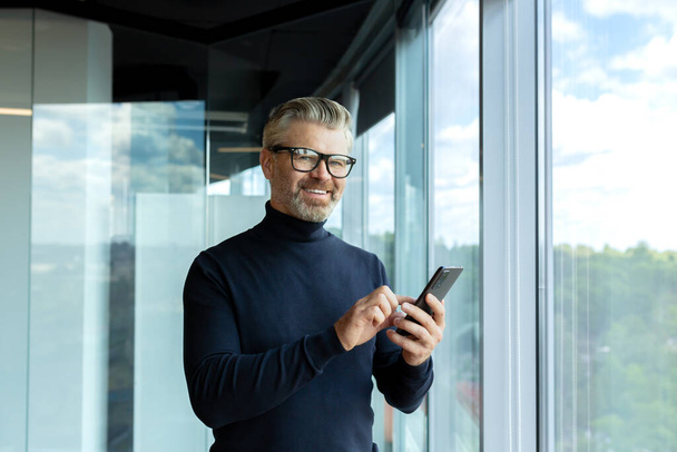 Portrait of senior handsome gray-haired businessman in glasses standing in skyscraper office near window, holding and uses a mobile phone, looks into the camera, smiles. - Photo, image