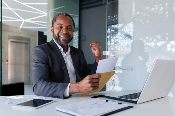 A senior African-American man in a suit is sitting at a desk in an office. Shows a joyful victory hand gesture. He reads the letter in the envelope. Got some good news, smiling at the camera. - 写真・画像
