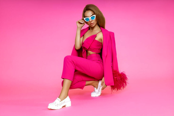 Elegant woman is wearing elegant pink jacket, trousers and white boots on pink background. Summer fashion style. Chic female model posing in stylish outfit. - Valokuva, kuva