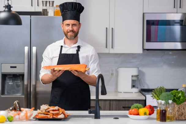 Millennial hispanic man in chef apron and cook hat hold salmon fillet at kitchen. Cooking salmon fillet. Restaurant menu with salmon fillet. Cooking, advertising salmon fillet concept - 写真・画像