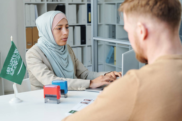 Modern Muslim woman working in Saudi Arabia embassy consulting unrecognizable man for visa application - Fotó, kép