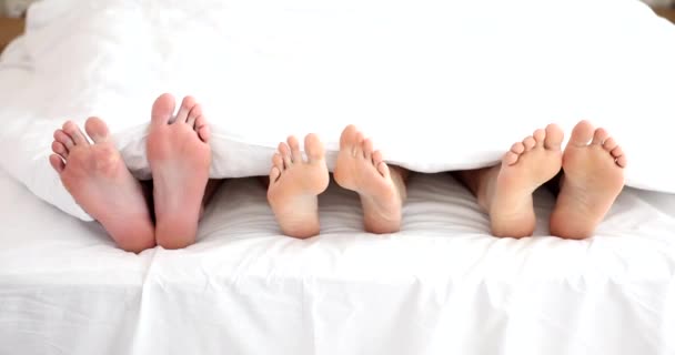 Grote voeten van ouders en klein kind bewegen bedekt met witte deken in de slaapkamer. Familieleden liggen op bed steken benen slow motion - Video