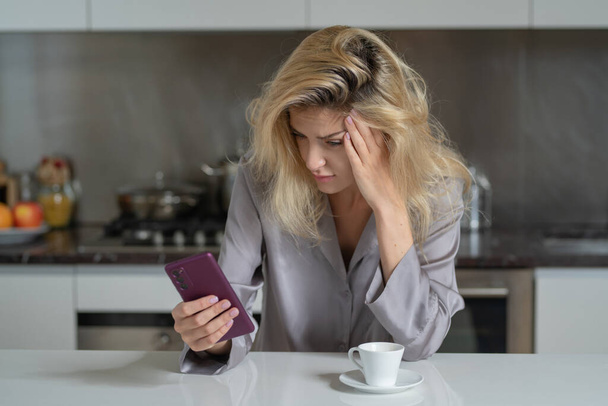Depressed woman in the kitchen in the morning. Upset girl with phone with coffee in kitchen. Morning of pretty woman. Sad lady using smartphone - Foto, imagen