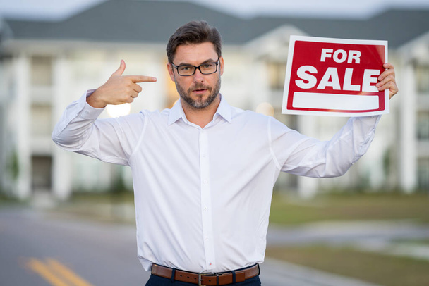 Realtor agent is a realtor with sign for sale in hand against the background on new apartment home background. Realtor in shirt, outdoor portrait. Realtor offering new home. Property concept - Foto, Bild