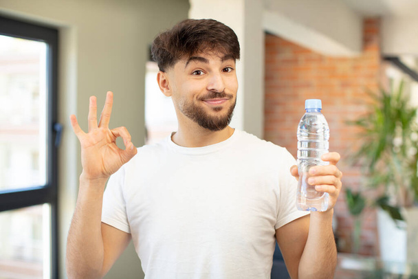 young handsome man feeling happy, showing approval with okay gesture. water bottle concept - Photo, Image