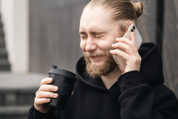 A serious young bearded uses smartphone and drinking coffee. Reusable eco friendly sustainable handy bamboo cup zero waste, no plastic concept. - Photo, Image