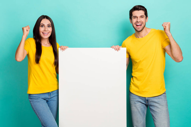 Photo of positive lucky husband wife dressed yellow t-shirts rising fist holding poster empty space isolated turquoise color background. - Valokuva, kuva