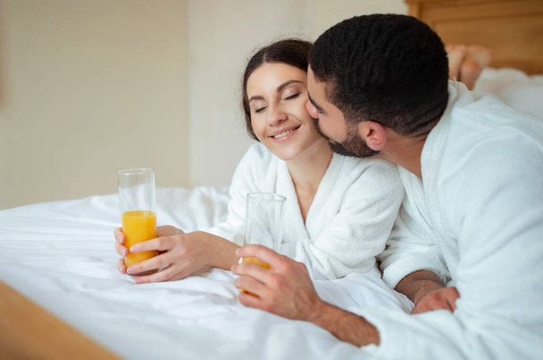 Vacanza romantica. Uomo baciare donna sdraiata a letto tenendo succo d'arancia indossare accappatoi bianchi in camera d'albergo moderna coperta. Multiculturale coppia relax a letto godendo luna di miele - Foto, immagini