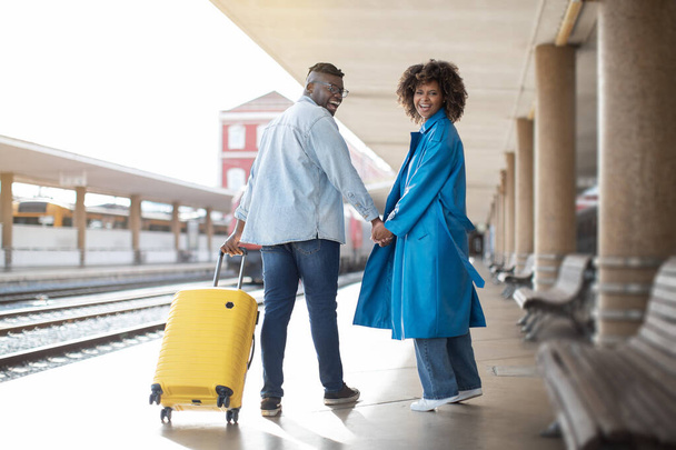 Portrait de couple noir romantique marchant avec une valise à la gare, heureux conjoints afro-américains tenant la main et tournant à la caméra tout en allant à train départ, copier l'espace - Photo, image