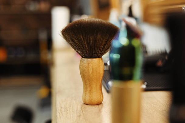 Fluffy retro black shaving brush with wooden handle on wooden work place table with blurred background of hair salon for men, barber shop. Selective focus. - Foto, imagen