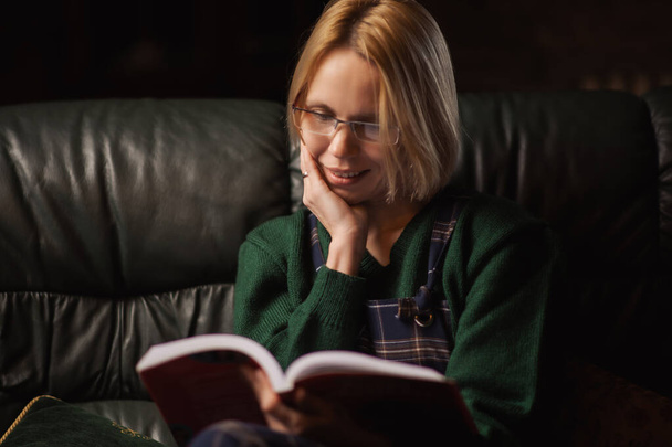 A woman with glasses reads a book on the sofa at home. Forty years old woman is reading a fascinating book. - Φωτογραφία, εικόνα