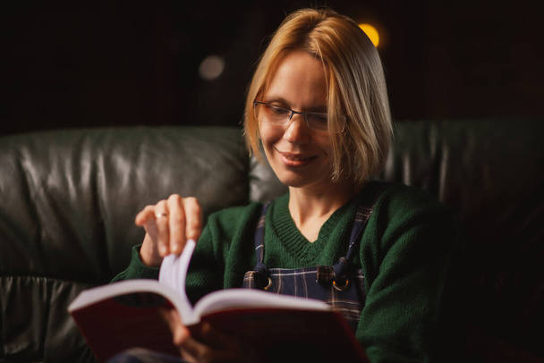 Uma mulher com óculos lê um livro no sofá em casa. Mulher de 40 anos está lendo um livro fascinante. - Foto, Imagem