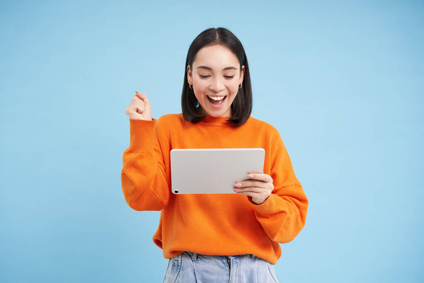 Joyeux jeune femme avec tablette numérique, regarde heureux gadget et célébrer, rire et sourire, debout sur fond bleu. - Photo, image