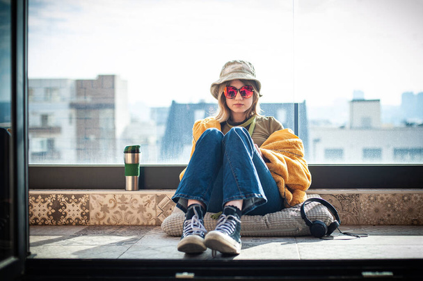 Copy space. Beautiful teenage girl, internet influencer sitting on patio floor on balcony, using smartphone, taking a selfie and entertainment. Yellow fur coat and red glasses. City in background. - Foto, imagen