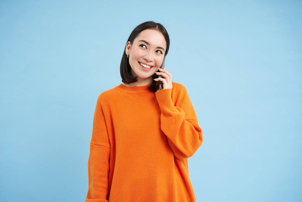 携帯電話で話すアジア系の若い女性の肖像画は、携帯電話を保持し、話す、青い背景の上に立つ. - 写真・画像