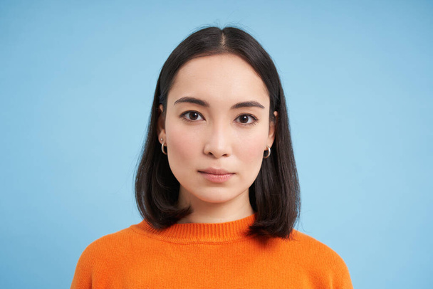 Primo piano di candida giovane donna asiatica, guardando la fotocamera con genuine emozioni naturali, in piedi su sfondo blu. - Foto, immagini