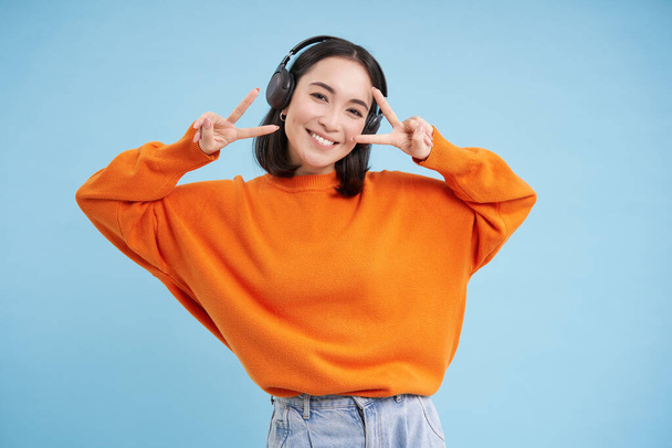 Beautiful korean woman in headphones, dancing and listening music in earphones, standing over blue background. - Foto, immagini
