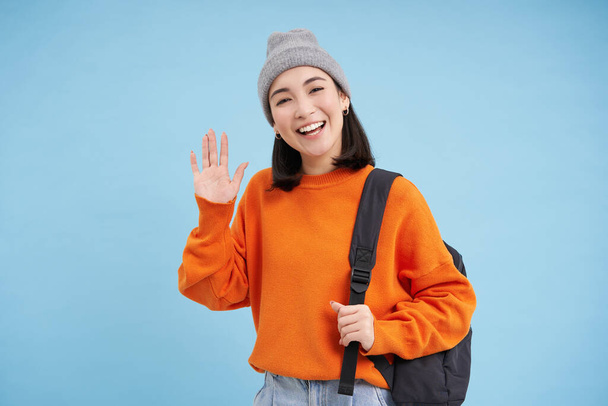 Friendly girl says hello, holds backpack and waves hand, greets you and laughs, stands over blue background. - Photo, Image