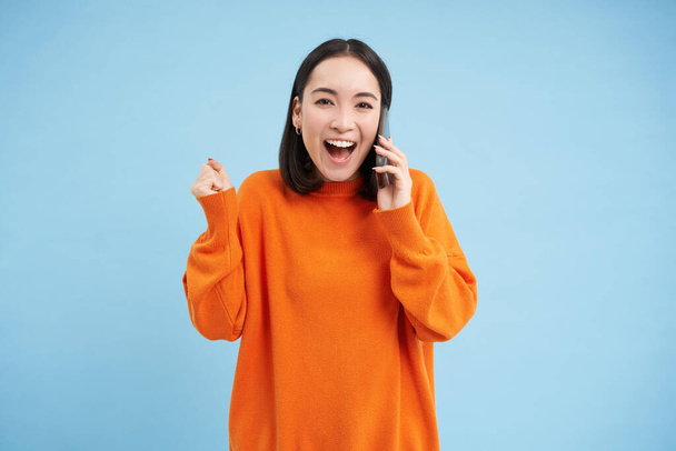 Mujer coreana sorprendida responde a la llamada telefónica y reacciona a las grandes noticias, celebrando, de pie sobre fondo de estudio azul. - Foto, imagen