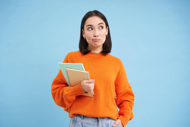 Přemýšlející dívka, student se zápisníky, stojí nad modrým pozadím přemýšlivý, rozhodování. - Fotografie, Obrázek